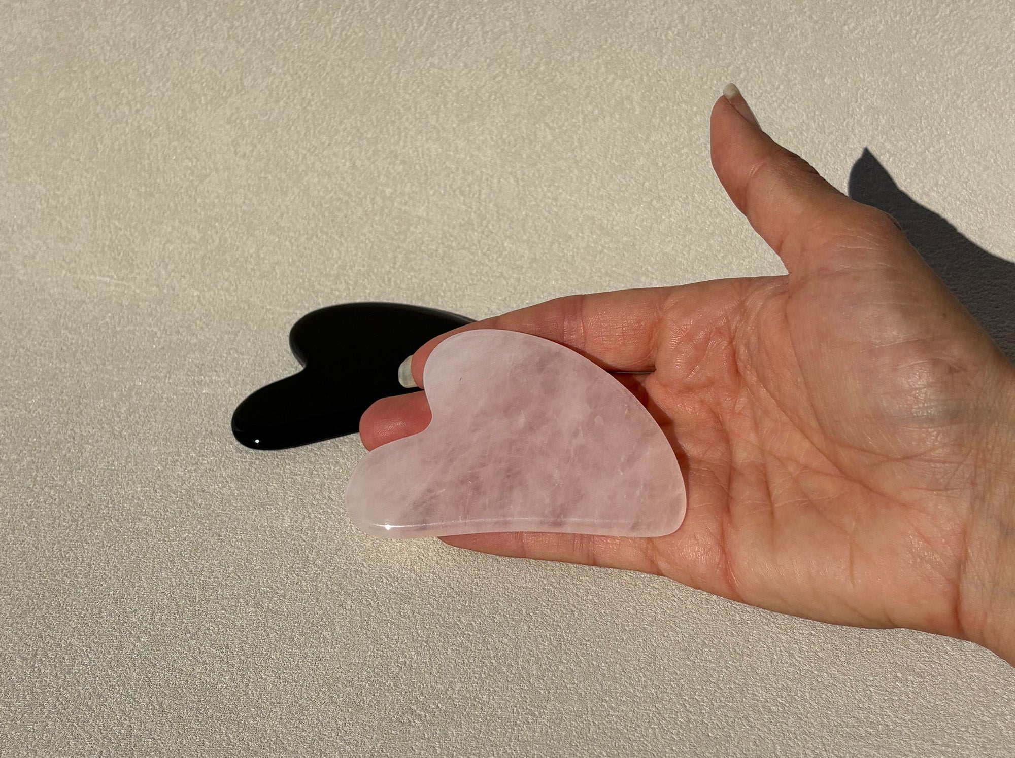 Eine Hand hält den Gua Sha Stein in der Hand um die Grösse zu zeigen. Die Steine sind aus schwarzem Obsidian und rosa Rosenquarz.