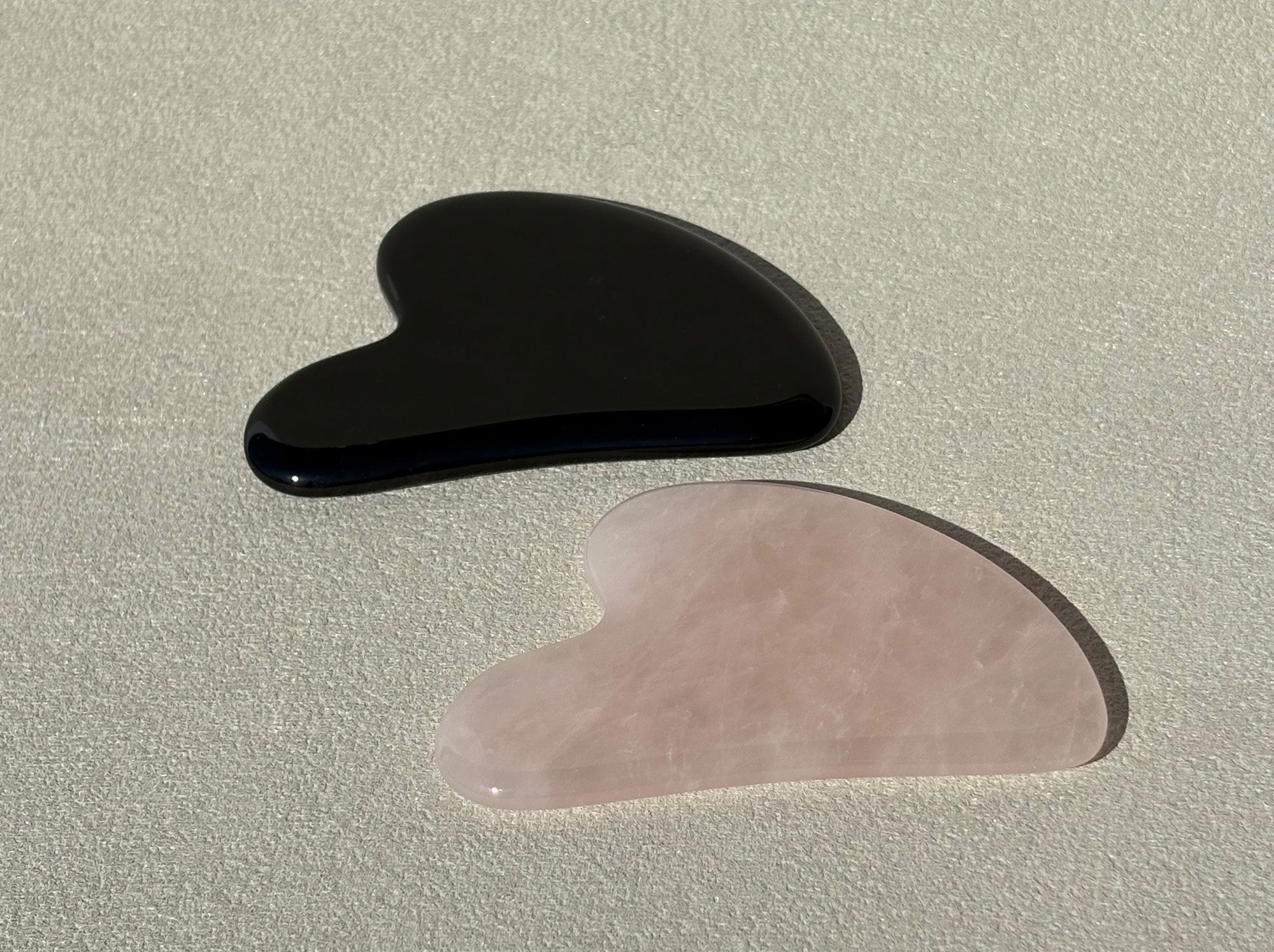 Zwei Gua Sha Steine liegen auf einer grauen Oberfläche, leicht aufgeraut. Die Steine sind aus schwarzem Obsidian und rosa Rosenquarz.