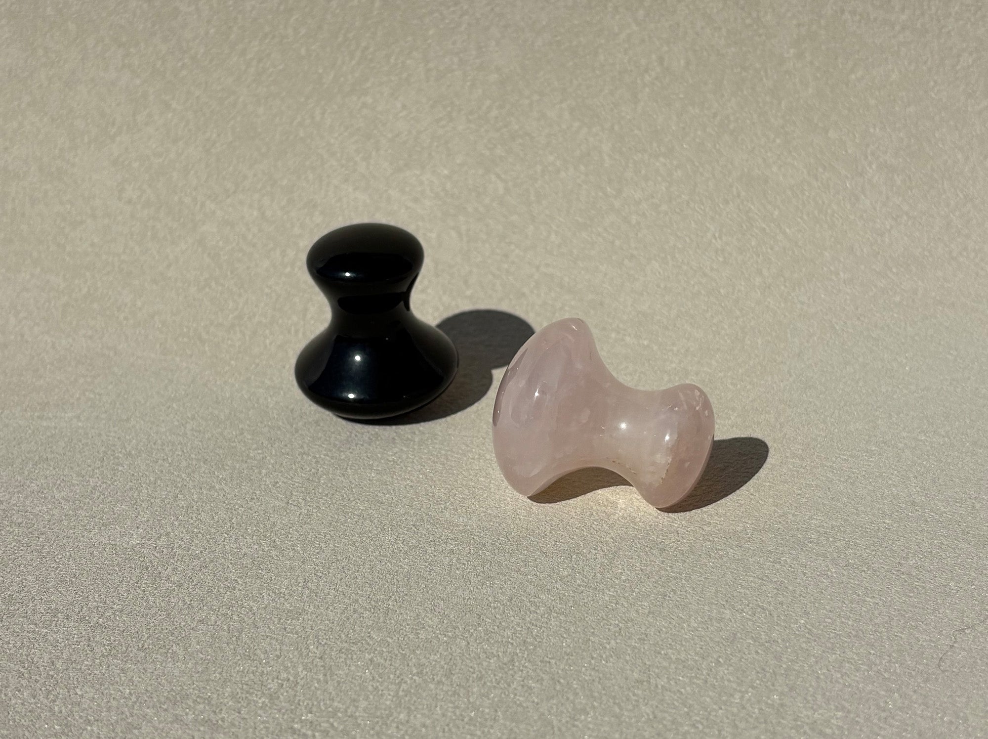 zwei Gua Sha Steine in Pilzform liegen auf einer grauen Oberfläche, leicht aufgeraut. Die Steine sind aus schwarzem Obsidian und rosa Rosenquarz.
