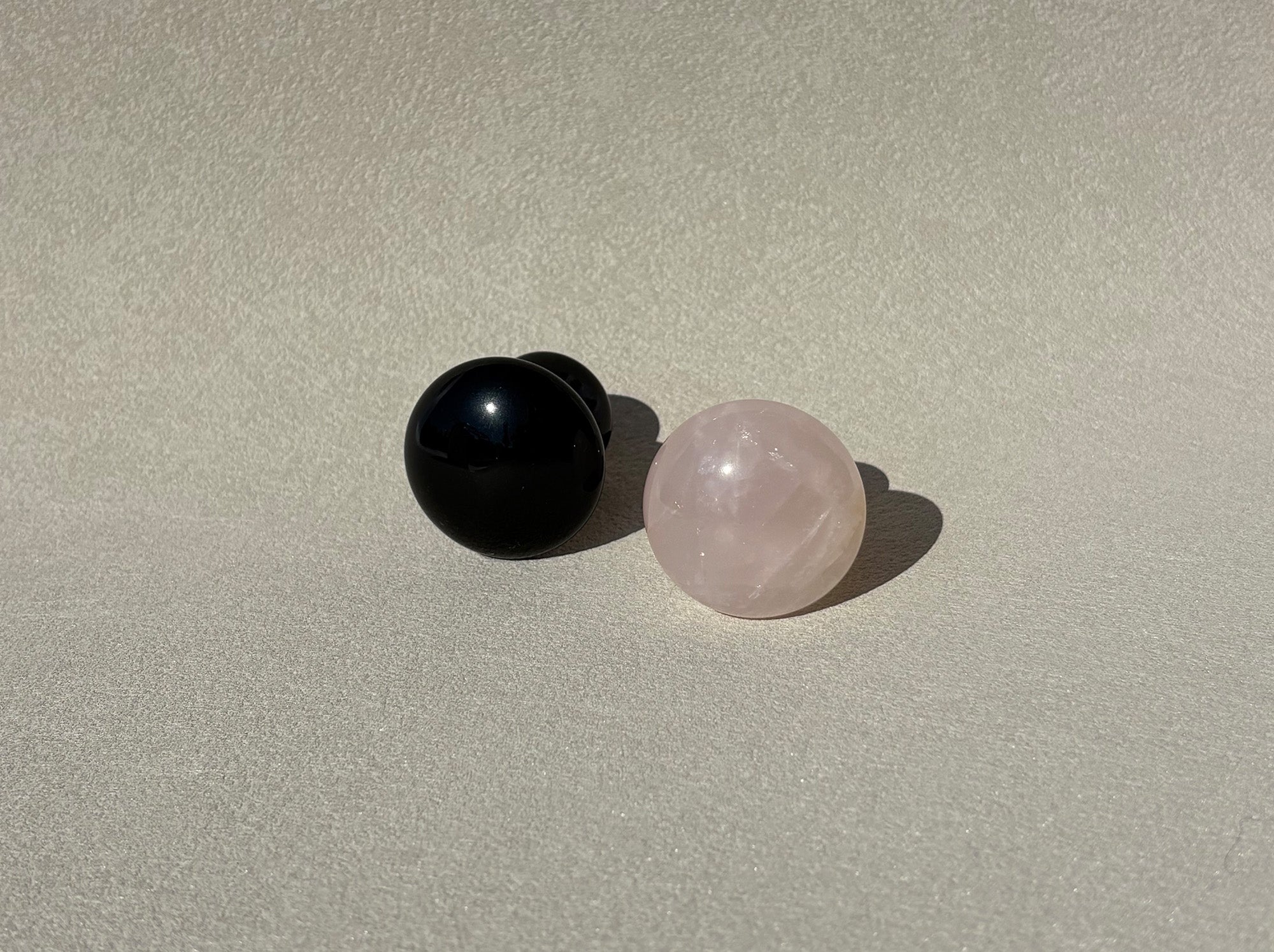 Die zwei Gua Sha Steine liegen auf einer grauen Oberfläche mit dem Kopf gegen die Kamera. Man sieht die runde Oberseite. Die Steine sind aus schwarzem Obsidian und rosa Rosenquarz.
