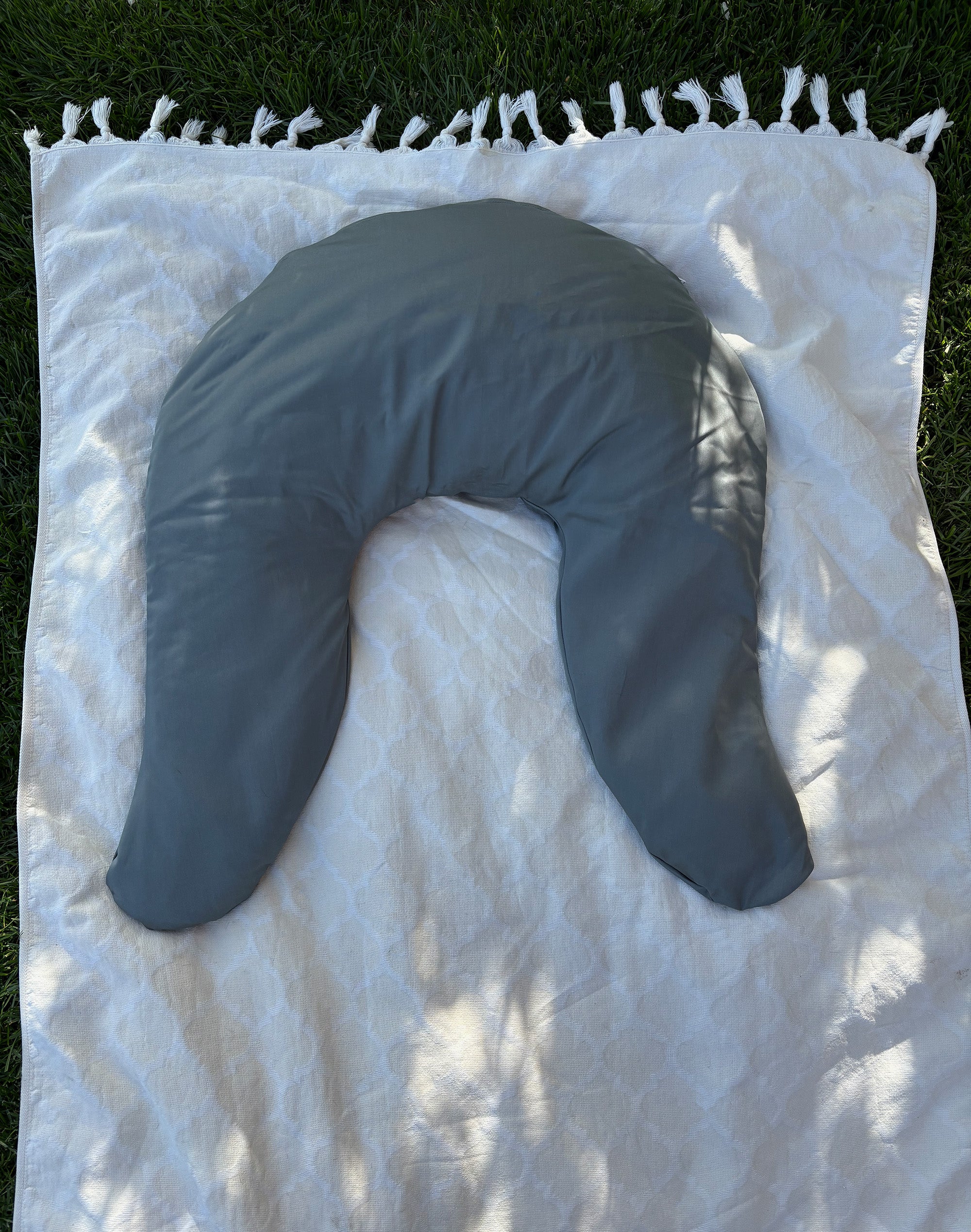 Das Spezialkissen Sana von SIELA liegt im Garten auf einem beigen Tuch. Der Rasen ist im Hintergrund zu sehen. Das Kissen ist mit einem Baumwoll-Satin Kissenbezug in der Farbe vert-gris zu sehen.